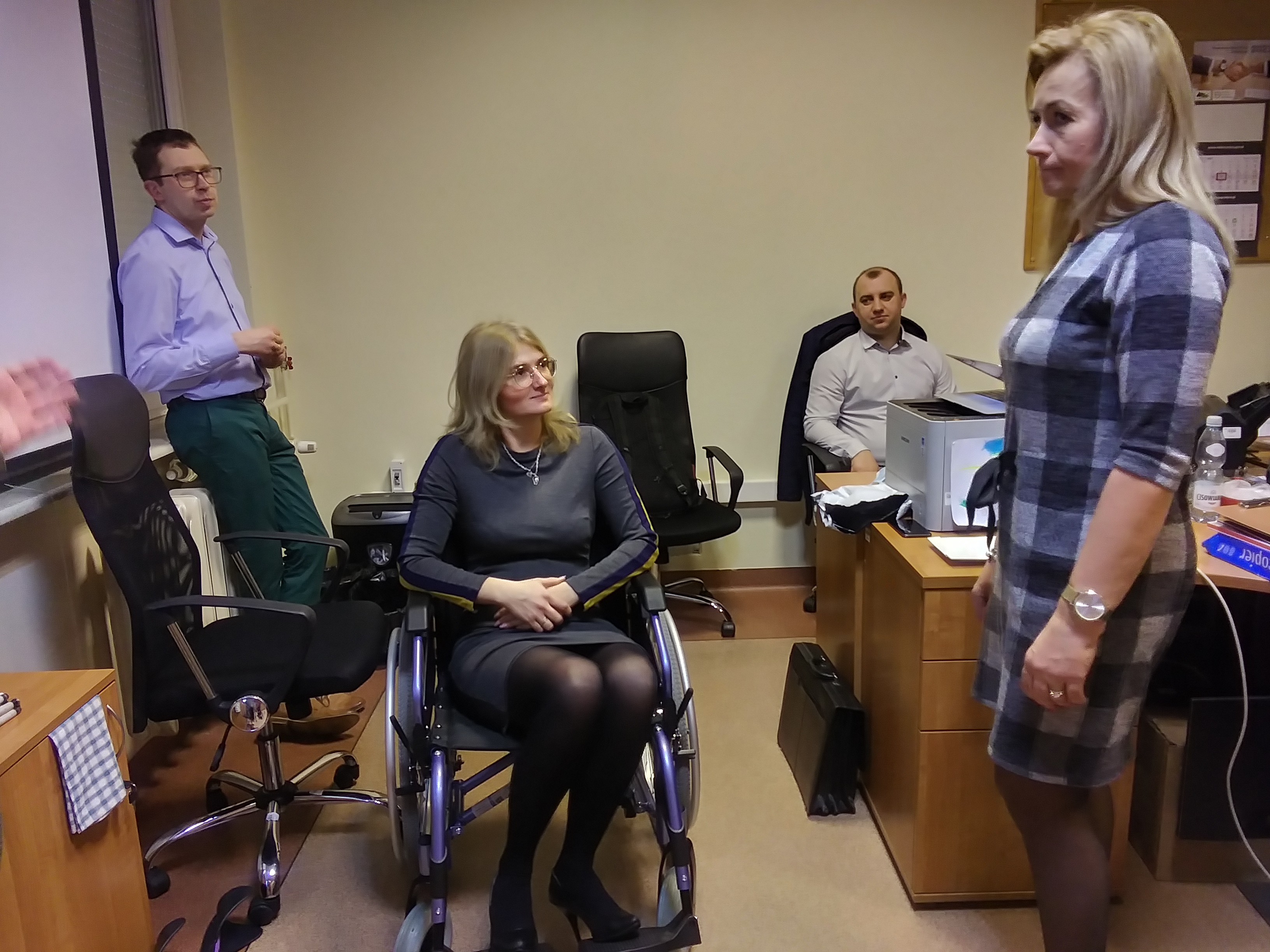 Zdjęcie przedstawia symulację niepełnosprawności ruchowej - pracownica PUP siedzi na wózku, obok niej widoczni są również inni uczestnicy szkolenia.