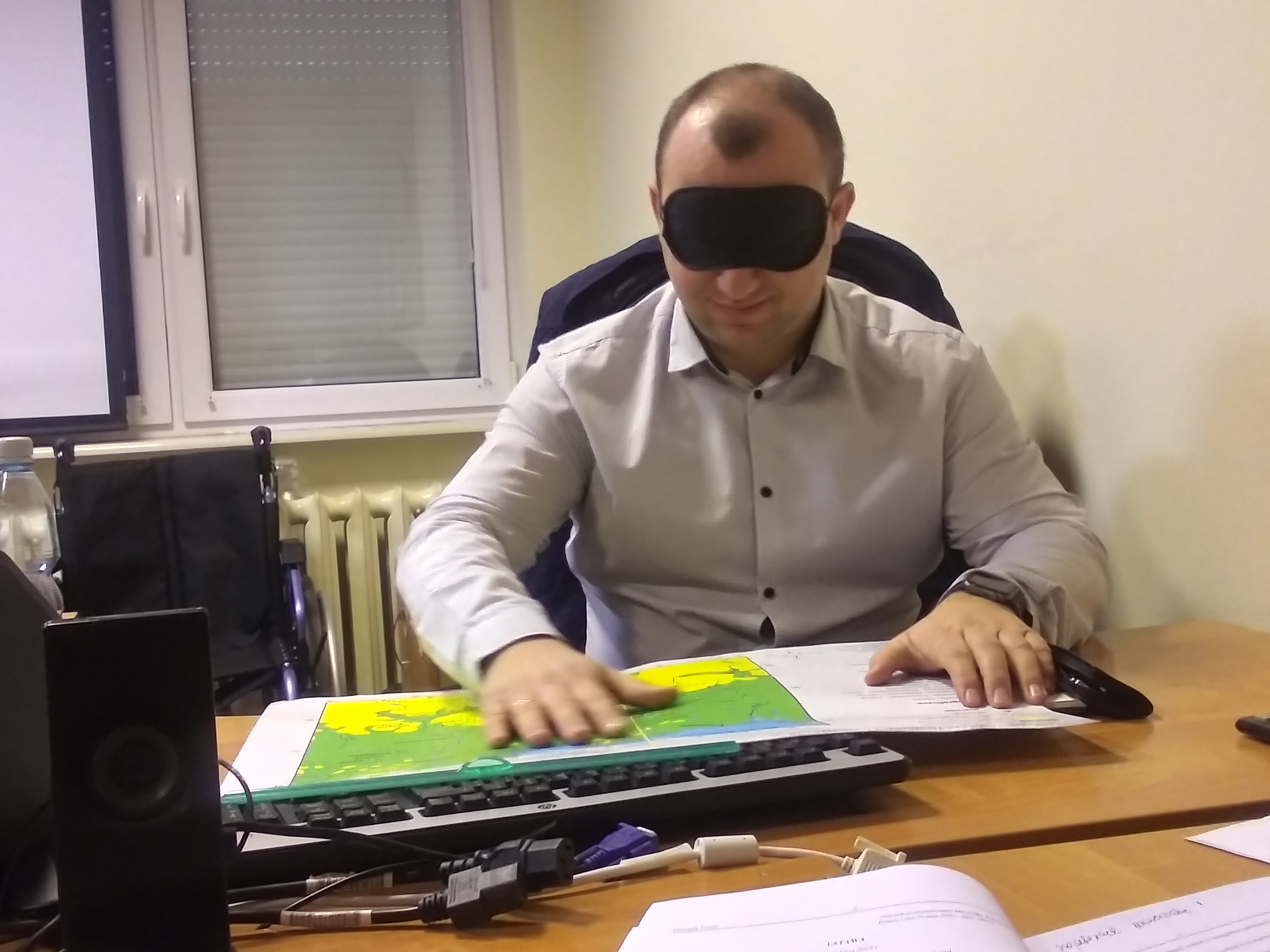 Zdjęcie przedstawia symulację niepełnosprawności wzrokowej - pracownik z zasłoniętymi oczami próbuje czytać mapę w piśmie Brajla